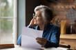 © fizkes - Thoughtful middle aged senior woman in eyeglasses holding paper document in hands, looking in distance. Pensive smiling older mature lady feeling pleased after reading correspondence letter at home.