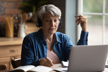 Focused middle aged mature woman wearing wireless headphones, involved in online communication with teacher using computer software video call application, older people and distant education concept.