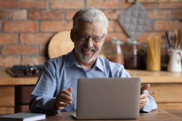 Happy older businessman in eyeglasses looking at laptop screen, feeling excited of reading email with good news. Laughing middle aged man enjoying online web camera video call talk with grown children