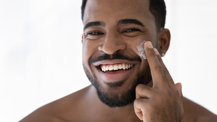 Wall Mural - Close up head shot happy laughing young 30s african american handsome man applying moisturizing cream on cheeks, feeling excited of using professional cosmetics in domestic skincare procedure.