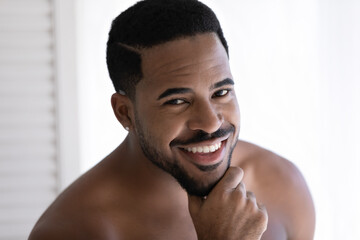 Wall Mural - Close up head shot handsome shirtless bare young 30s african american man touching unshaved chin, looking at camera, posing for photo, feeling confident alone indoors, masculine mixed race beauty.