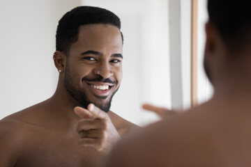 Mirror reflection close up head shot naked smiling 30s young african ethnicity man pointing finger looking at camera, enjoying starting energetic day after morning cleaning routine in bathroom.