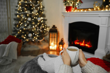 Poster - Woman with hot drink resting near fireplace in cozy room decorated for Christmas, closeup