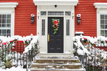 Snow Covered Residential Stairs Leading To A Black Door With White Wood Trim And Windows. The Stair Rails Have Fresh Evergreen Garlands With Bright Red Bows. A Christmas Wreath Hangs On The Door. 