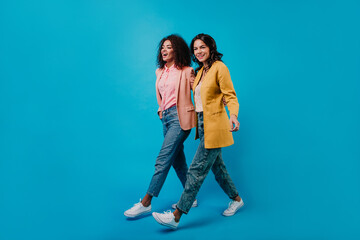 Full-length studio shot of two trendy girls walking on blue background. Smiling african and latin women posing in jeans.