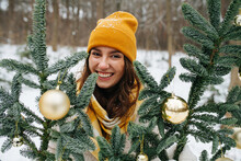Happy Smiling Girl Outside At Winter Behind Synthetic Fir Tree Decorated With Shiny Christmas Balls. She Wears Yellow Knit Scarf And Hat.