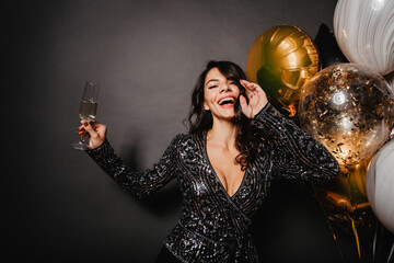 Wall Mural - Slim latin woman having fun at event. Debonair girl with glass of wine dancing on black background.