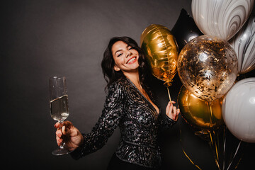 Pleased latin girl standing near party balloons. Enchanting young woman drinking wine in her birthday.