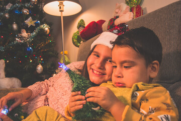 Escena infantil navideña. Niño y niña sentados en el sofá con el árbol de Navidad detrás. Infancia feliz de dos hermanos.