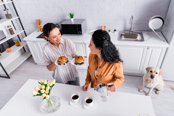 High angle view of smiling hispanic lesbian woman with pancakes looking at partner  during breakfast in kitchen