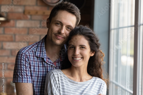 Strong marriage. Headshot portrait of happy affectionate young spouses standing at home by large window. Couple in love bonding smiling looking at camera demonstrating harmony romance in relations