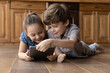 © fizkes - Cozy leisure with gadget. Happy brother sister of primary school age lying on warm floor with heating system at kitchen using pad pc. Smiling little kids play game on tablet watching video having fun