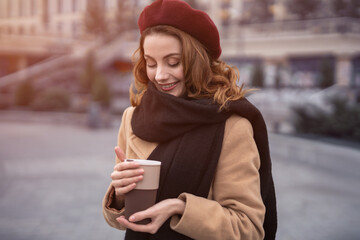 Half-length portrait of a beautiful french styled young woman holding a coffee mug standing outdoors. Happy french stylish young woman wearing autumn coat and red beret. Tinted image. 