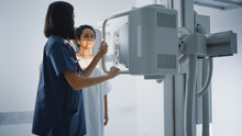 Hospital Radiology Room: Beautiful Latin Woman Standing While Female Radiologist Adjusts X-Ray Machine. Preventive Treatment Scanning Chest, Back, Breast, Lungs. Modern Clinic With Advanced Technology