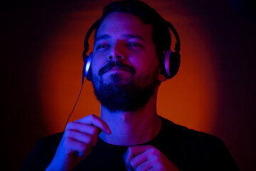 Young man headshot while keeping the rhythm of the music listening on headphones. He is illuminated with red and blue lights and there is a orange spot light in the background.