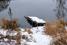 A Broken Bridge Covered With Snow On The Bank Of A River With Dry Yellow Grass.