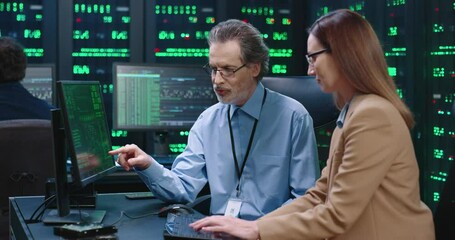 Senior employee of system administrator with glasses sits at table with female intern programmer and explains to her workflows showing on computer screen.