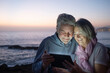 © luciano - Cheerful senior couple white-haired in video chat with a digital tablet, sitting in the dusk on the beach with horizon over water on background. Happy retirement concept together