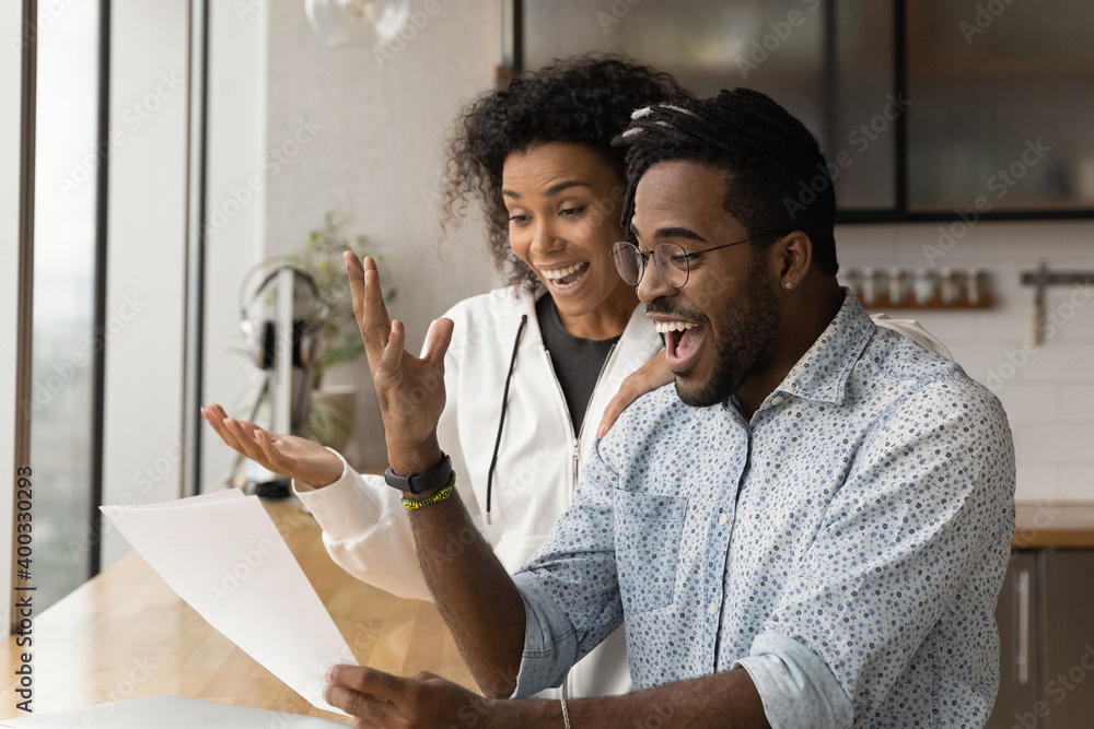 Happy young African American couple feel euphoric read good unbelievable news in paper letter correspondence. Overjoyed biracial man and woman triumph get pleasant message in paperwork. - obrazy, fototapety, plakaty 