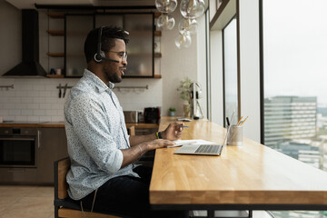 Wall Mural - Young biracial man in headphones talk on video call on laptop in home office, make notes in notebook. African American male employee in earphones have webcam digital virtual conference online.