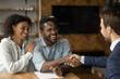 Happy African American couple shake hand close deal with male realtor or broker at meeting. Smiling biracial young family man and woman handshake make agreement with real estate agent in office.