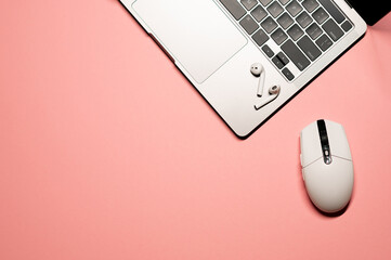 Minimalistic flat lay photo on a pastel pink background with a modern silver grey laptop, wireless earphones, and a white plastic computer mouse.