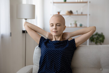 Wall Mural - Calm sick millennial female relax nap on cozy sofa at living room breath fresh air hold hands behind head. Serene young hairless woman diagnosed with oncology rest on couch dream enjoy peace of mind