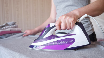 Hardworking woman holding modern purple electric iron irons fast and carefully grey shirt after laundry on special board at home extreme closeup