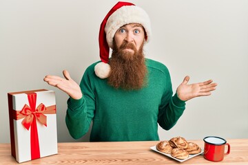 Wall Mural - Young irish redhead man wearing christmas sitting on the table with gift clueless and confused expression with arms and hands raised. doubt concept.