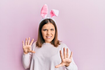 Sticker - Young beautiful woman wearing cute easter bunny ears afraid and terrified with fear expression stop gesture with hands, shouting in shock. panic concept.