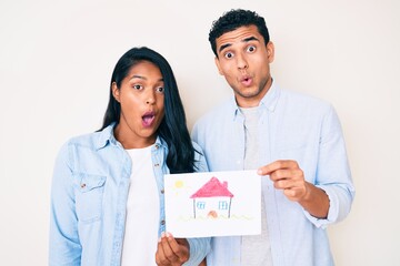 Canvas Print - Beautiful latin young couple showing house proyect on a paper scared and amazed with open mouth for surprise, disbelief face