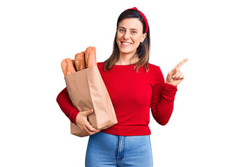 Wall Mural - Young beautiful woman holding paper bag with bread smiling happy pointing with hand and finger to the side