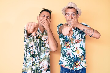 Wall Mural - Young gay couple of two men wearing summer hat and hawaiian shirt laughing at you, pointing finger to the camera with hand over mouth, shame expression