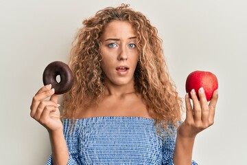 Sticker - Beautiful caucasian teenager girl holding red apple and donut in shock face, looking skeptical and sarcastic, surprised with open mouth