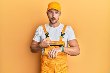 Canvas Print - Young handsome man wearing handyman uniform over yellow background in hurry pointing to watch time, impatience, upset and angry for deadline delay