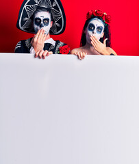 Wall Mural - Young couple wearing mexican day of the dead costume holding blank empty banner covering mouth with hand, shocked and afraid for mistake. surprised expression