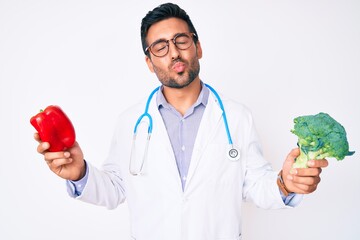 Young hispanic man wearing doctor stethoscope holding red pepper and broccoli looking at the camera blowing a kiss being lovely and sexy. love expression.