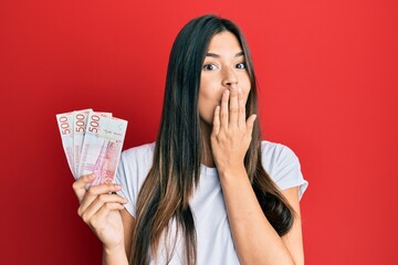 Wall Mural - Young brunette woman holding 500 swedish krona banknotes covering mouth with hand, shocked and afraid for mistake. surprised expression