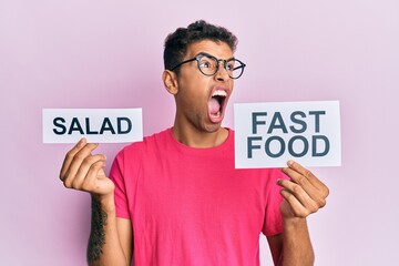 Sticker - Young handsome african american man holding salad and fast food message paper angry and mad screaming frustrated and furious, shouting with anger. rage and aggressive concept.