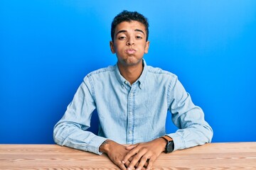 Poster - Young handsome african american man wearing casual clothes sitting on the table puffing cheeks with funny face. mouth inflated with air, crazy expression.