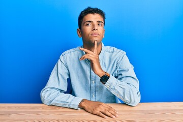 Sticker - Young handsome african american man wearing casual clothes sitting on the table thinking concentrated about doubt with finger on chin and looking up wondering