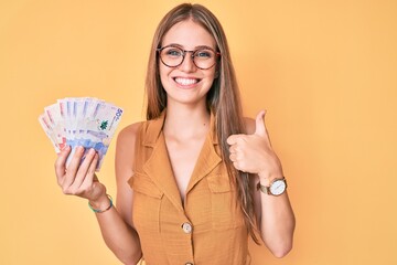 Wall Mural - Young blonde girl holding colombian pesos smiling happy and positive, thumb up doing excellent and approval sign