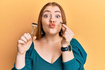 Sticker - Young beautiful redhead woman holding eyelash curler and fake lashes looking at the camera blowing a kiss being lovely and sexy. love expression.