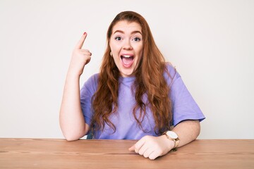 Poster - Young redhead woman wearing casual clothes sitting on the table pointing finger up with successful idea. exited and happy. number one.