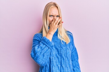 Sticker - Young blonde girl wearing wool winter sweater smelling something stinky and disgusting, intolerable smell, holding breath with fingers on nose. bad smell