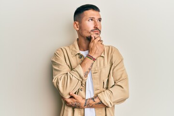 Wall Mural - Handsome man with tattoos wearing casual brown denim jacket with hand on chin thinking about question, pensive expression. smiling with thoughtful face. doubt concept.