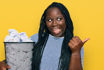 Sticker - Young black woman with braids holding paper bin full of crumpled papers pointing thumb up to the side smiling happy with open mouth