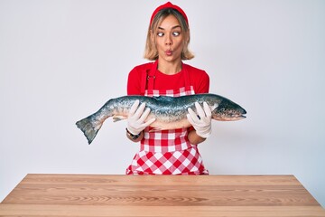 Beautiful caucasian woman fishmonger selling fresh raw salmon making fish face with mouth and squinting eyes, crazy and comical.