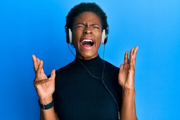 Canvas Print - Young african american girl listening to music using headphones celebrating mad and crazy for success with arms raised and closed eyes screaming excited. winner concept