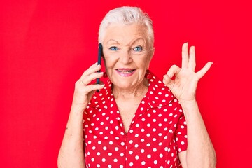 Senior beautiful woman with blue eyes and grey hair having conversation talking on the smartphone doing ok sign with fingers, smiling friendly gesturing excellent symbol